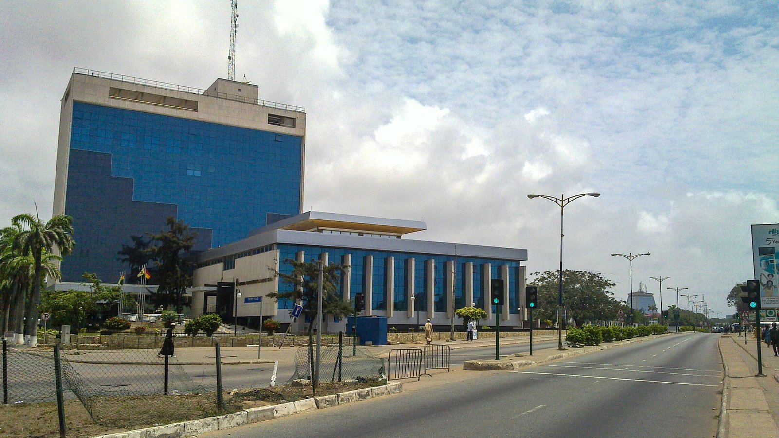 An Accra street beside a blue-glass building, with a tree-lined central divider and pedestrians on the pavement.