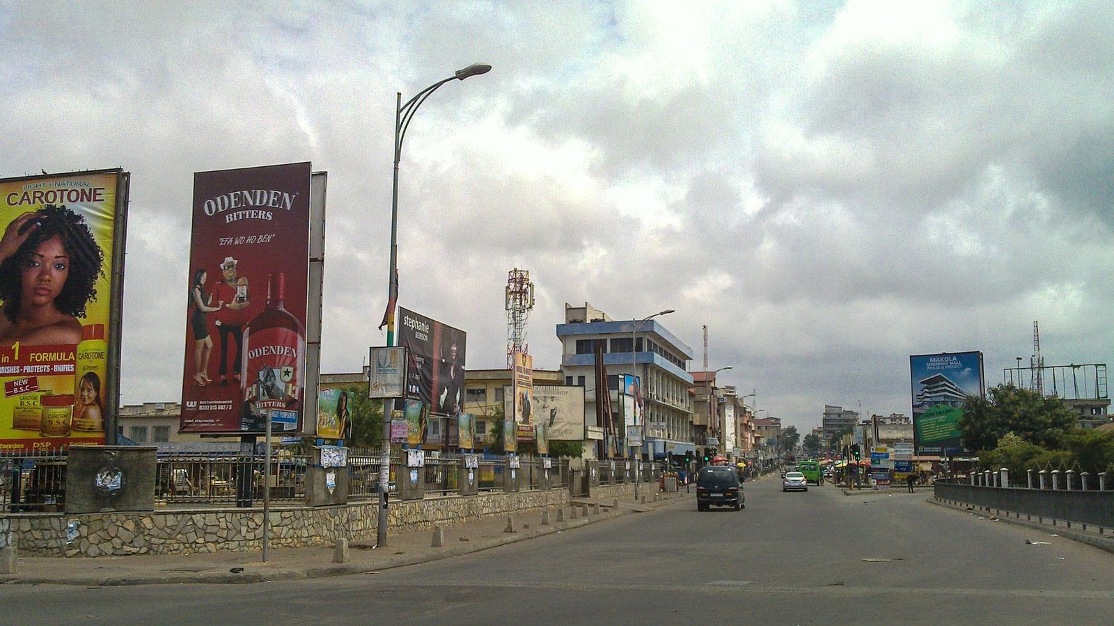 A wide road in Accra with roadside buildings, signs and light traffic under a cloudy sky.